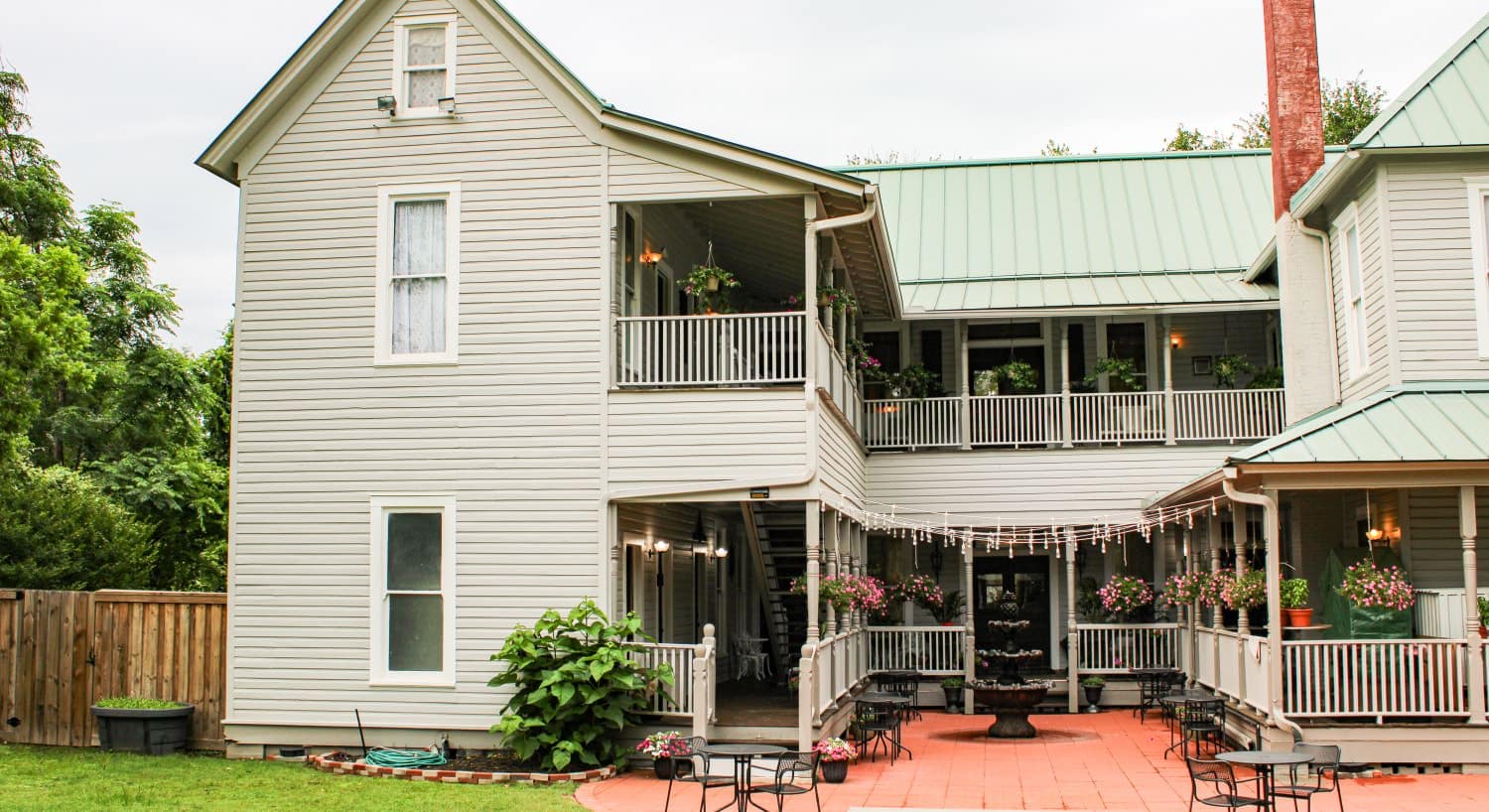 Exterior view of back of property painted light gray with white and dark gray trim surrounded by green grass and large trees and a red-bricked center patio with black iron patio tables and chairs