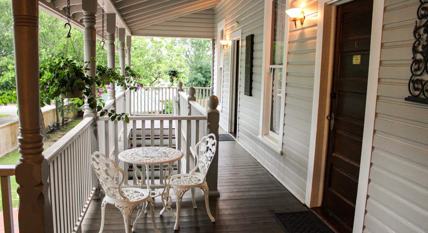 Upper level covered deck with white wrought iron table and chairs, hanging flower baskets, and dark wooden entry doors to rooms