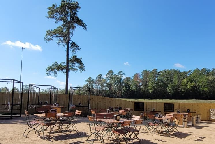 Large outdoor venue with patio tables and chairs surrounded by a wood fence with large green trees in the background