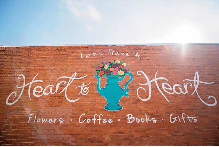 Mural of a flower-filled teapot and white text stating Let's Have A Heart to Heart Flowers, Coffee, Books, and Gifts on red brick wall