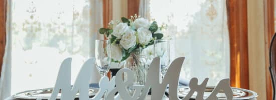 A round table with a dark blue linen tablecloth set with 2 silver charges, 2 champagne flutes, a vase with white roses, and white signs in front that say Mr & Mrs