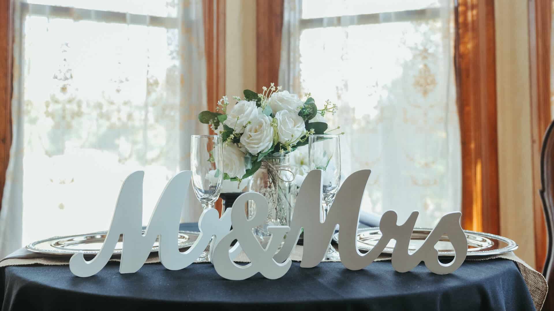 A round table with a dark blue linen tablecloth set with 2 silver charges, 2 champagne flutes, a vase with white roses, and white signs in front that say Mr & Mrs