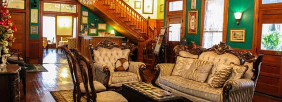 A living room with dark wood floors, green walls, antique furniture, an open doorway into a dining room, and a wood staircase leading to a 2nd floor.