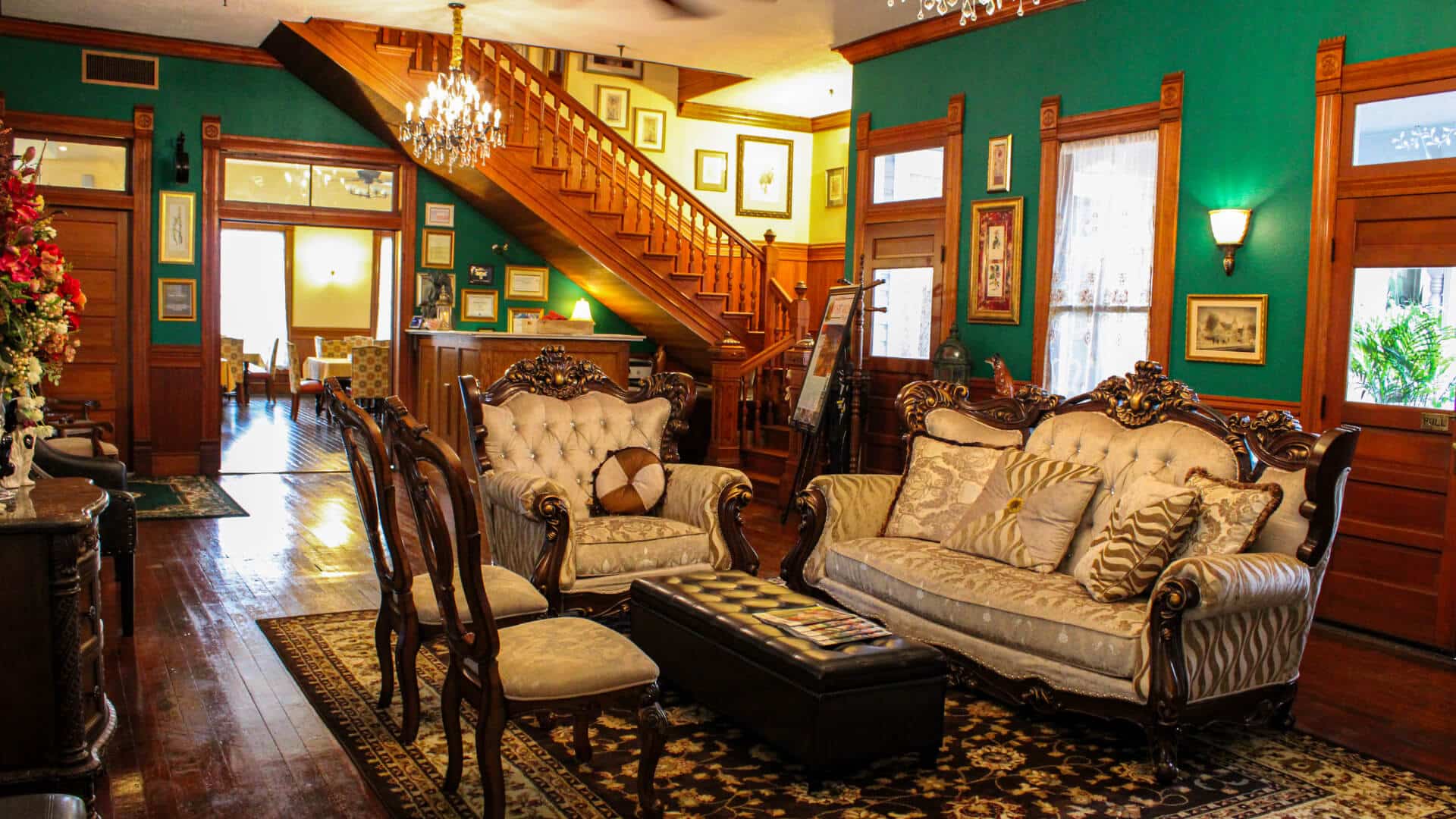 A living room with dark wood floors, green walls, antique furniture, an open doorway into a dining room, and a wood staircase leading to a 2nd floor.