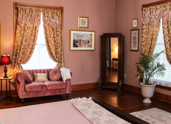 Bedroom with light pink walls, hardwood flooring, vintage floor mirror, and antique pink cushioned love seat