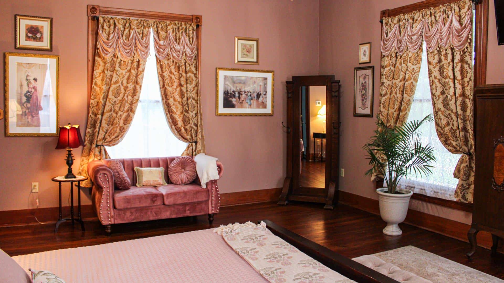 Bedroom with light pink walls, hardwood flooring, vintage floor mirror, and antique pink cushioned love seat