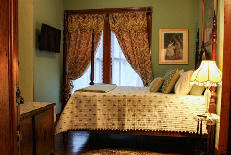 Bedroom with green walls, hardwood flooring, dark wooden antique four poster bed, white bedding, and antique wooden cabinet