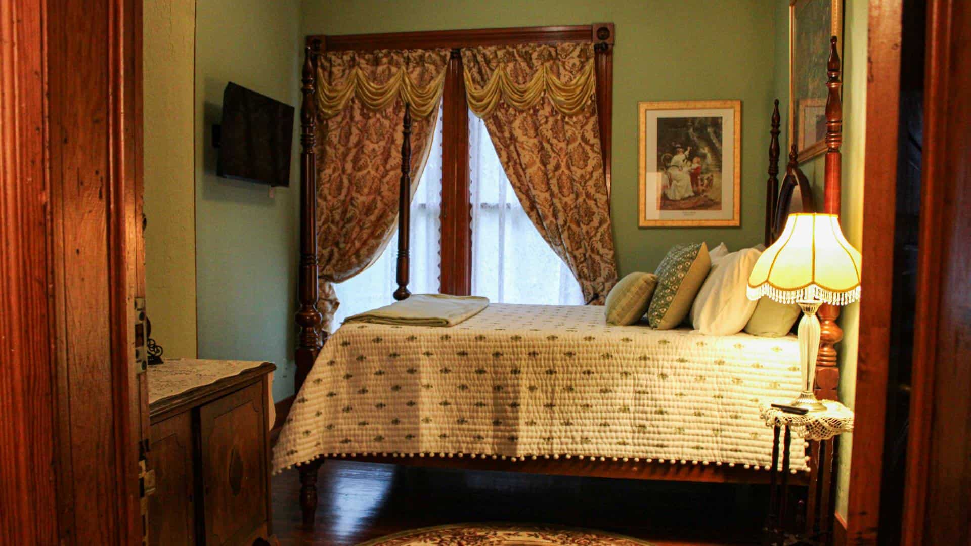 Bedroom with green walls, hardwood flooring, dark wooden antique four poster bed, white bedding, and antique wooden cabinet