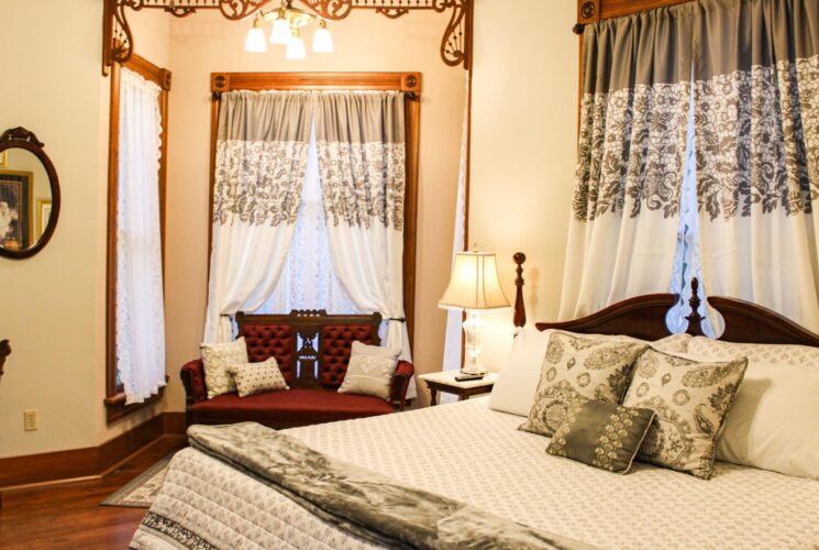 Bedroom with light-colored walls, hardwood flooring, antique dark wooden bed, white bedding, and antique loveseat