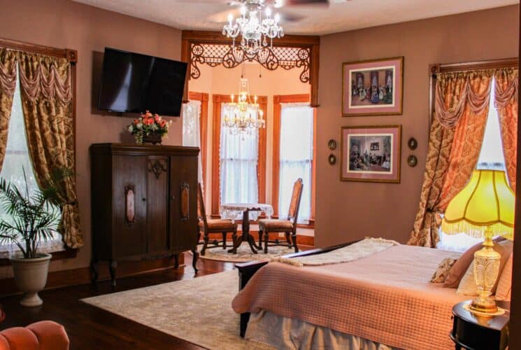 Bedroom with light pink walls, hardwood flooring, dark wooden bed, pink comforter, vintage wooden cabinet, and wall-mounted TV