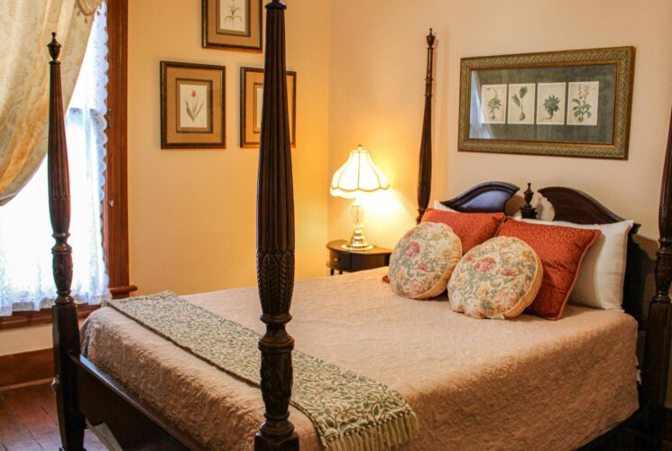 Bedroom with light-colored walls, hardwood flooring, dark wooden four-poster bed, light pink bedding, and antique dark wooden nightstand with lamp