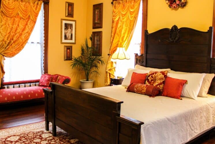 Bedroom with yellow walls, hardwood flooring, dark wooden antique bed, white bedding, and antique chaise lounge