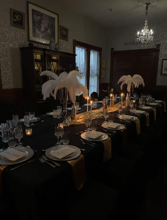 Dining room lighted up with candles and a chandelier and a long table in the middle with black tablecloth, gold accents, and multiple place settings for a dinner party