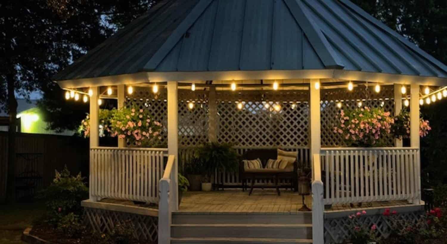 Backyard gazebo at night with hanging lights, hanging baskets, and patio furniture