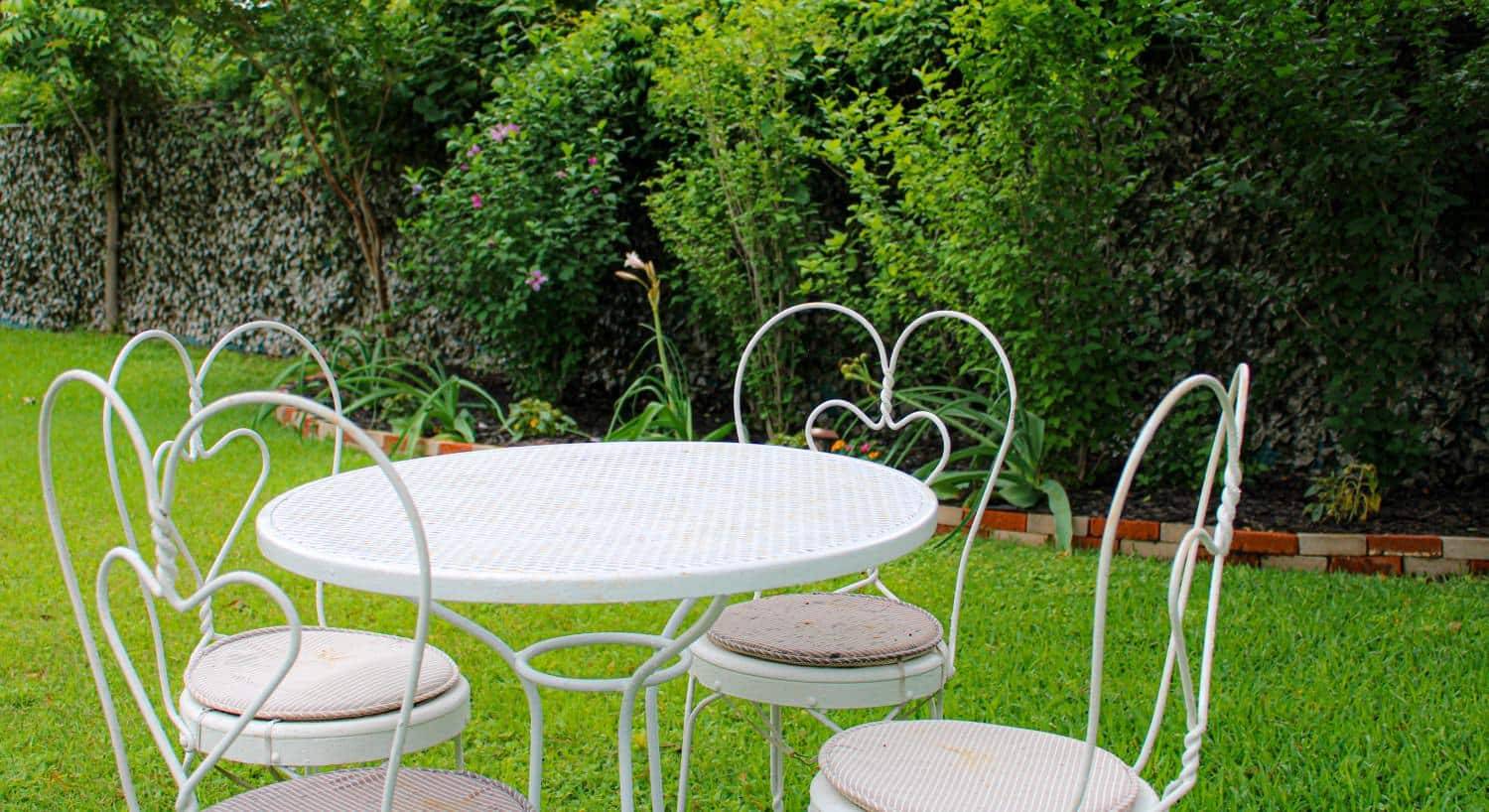 White metal patio table and chairs near green grass with green bushes in the back